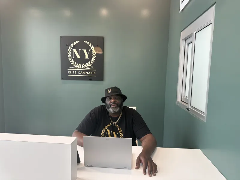 Carson Grant, founder of NY Elite Cannabis, seated beneath the gold NY Elite Cannabis laurel logo at the dispensary